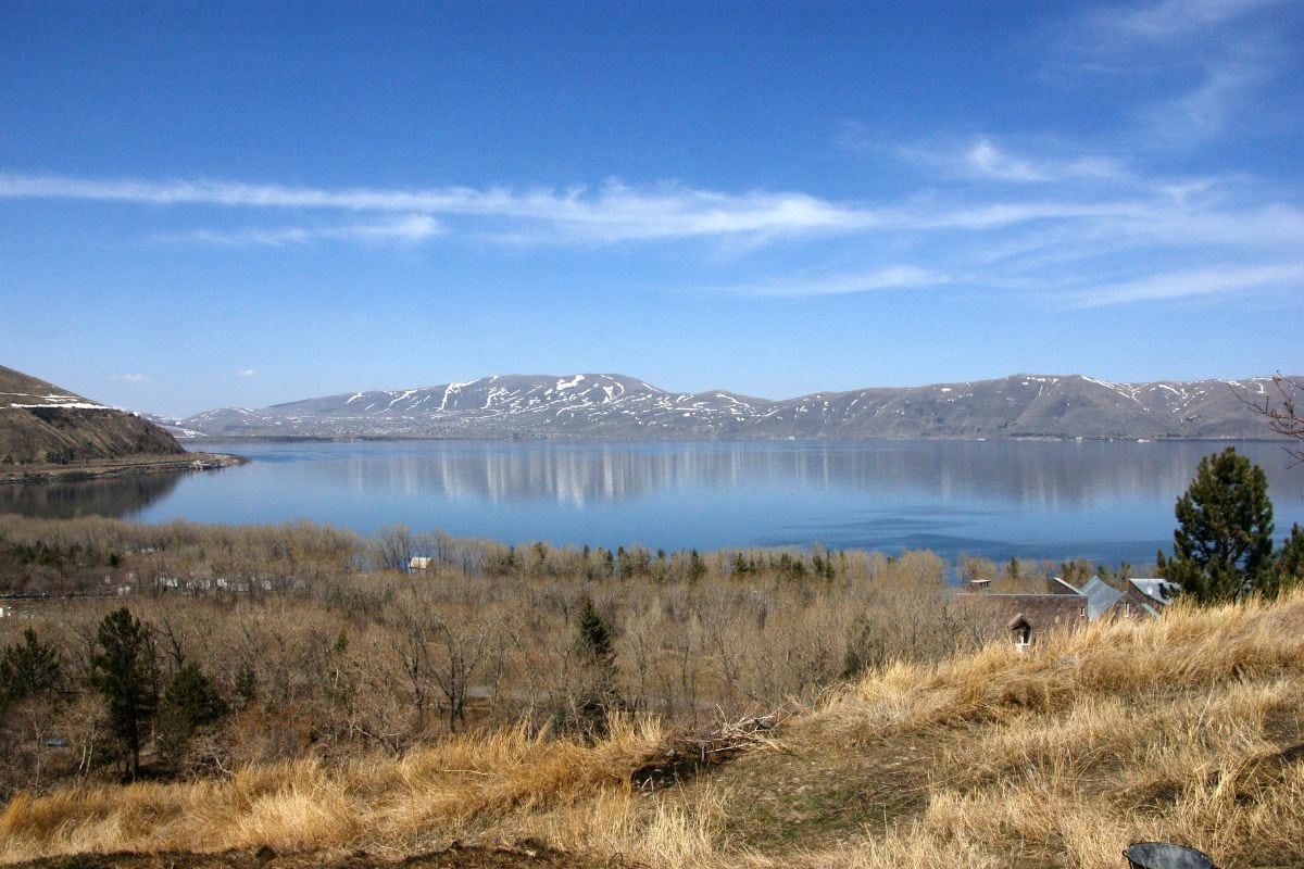 Blick auf einen ruhigen See mit schneebedeckten Bergen im Hintergrund und bewaldeten Ufern, unter einem klaren blauen Himmel.