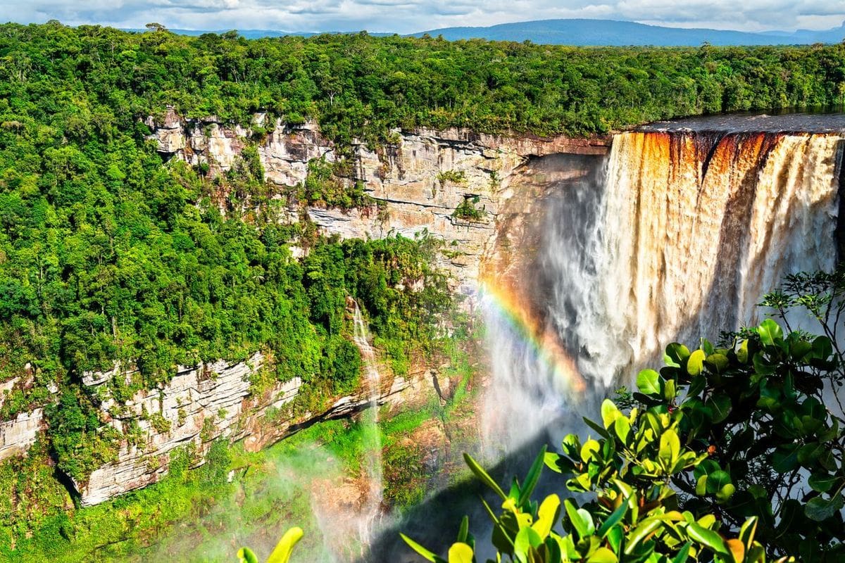 Ein beeindruckender Wasserfall umgeben von üppigem Grün und einem Regenbogen, der sich im Wassernebel bildet.