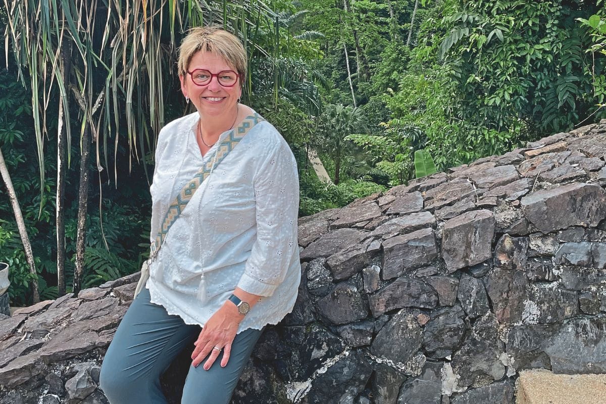 Eine Frau mit Kurzhaarschnitt und roter Brille trägt eine weiße Bluse und sitzt auf einer niedrigen Natursteinmauer vor einer üppigen, grünen Waldkulisse.