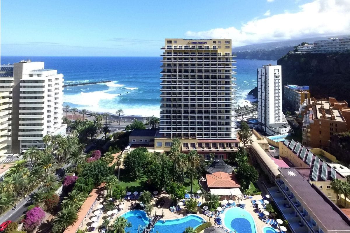 Blick auf ein Hotelgebäude mit Poolanlage und Palmen, umgeben von Küstenlandschaft und dem Meer im Hintergrund.
