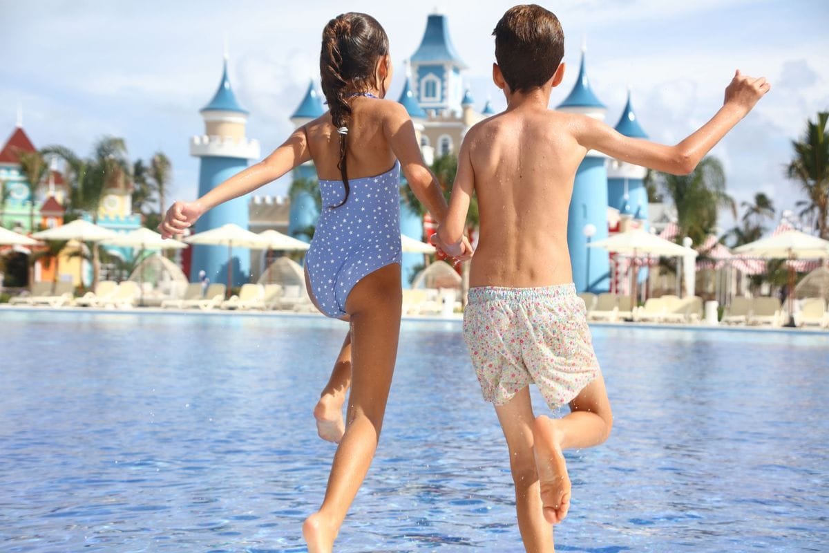 Ein Mädchen und ein Junge springen Hand in Hand in einen Swimmingpool vor einer Kulisse mit märchenhaften Türmen und Sonnenschirmen im Hintergrund.