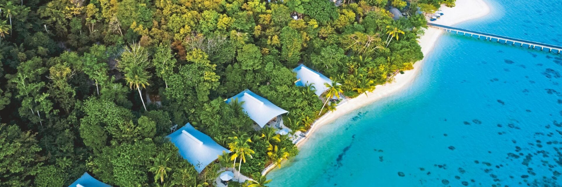 bawah reserve hero Luftaufnahme einer tropischen Küste mit weißem Sandstrand, dichtem Wald, mehreren Villen mit hellen Dächern und einem langen Steg im türkisfarbenen Wasser.