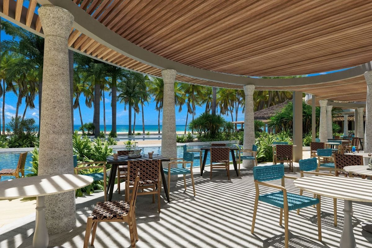 Eine überdachte Terrasse mit runden Steinsäulen und Holzdach, bestuhlt mit Tischen und Stühlen, bietet einen Blick auf einen Pool und Palmen am Sandstrand.