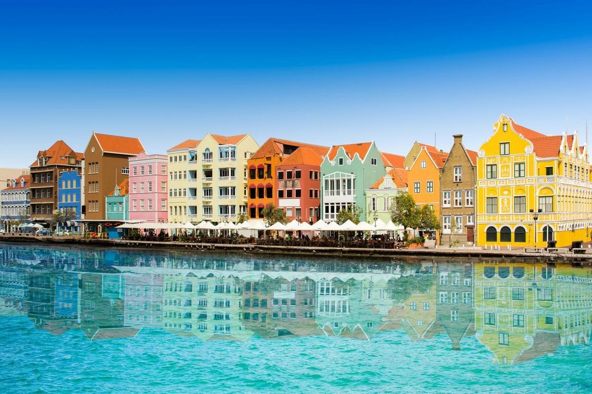 Eine Reihe von bunten, historischen Kolonialbauten in Willemstad spiegelt sich bei strahlend blauem Himmel klar im Wasser der angrenzenden Hafenbucht wider.