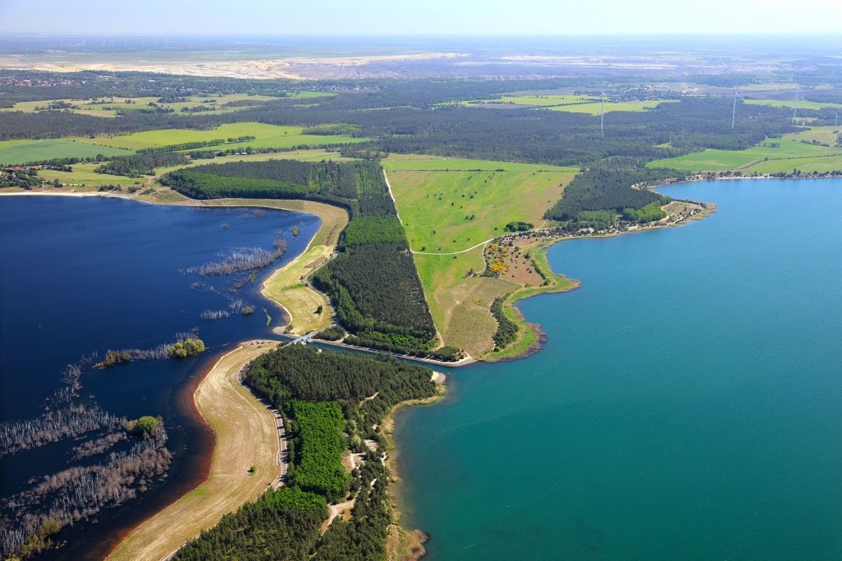 Luftaufnahme einer Seenlandschaft mit einem schmalen Landstreifen, der zwei unterschiedlich gefärbte Gewässer trennt, umgeben von Wald und Wiesenflächen.