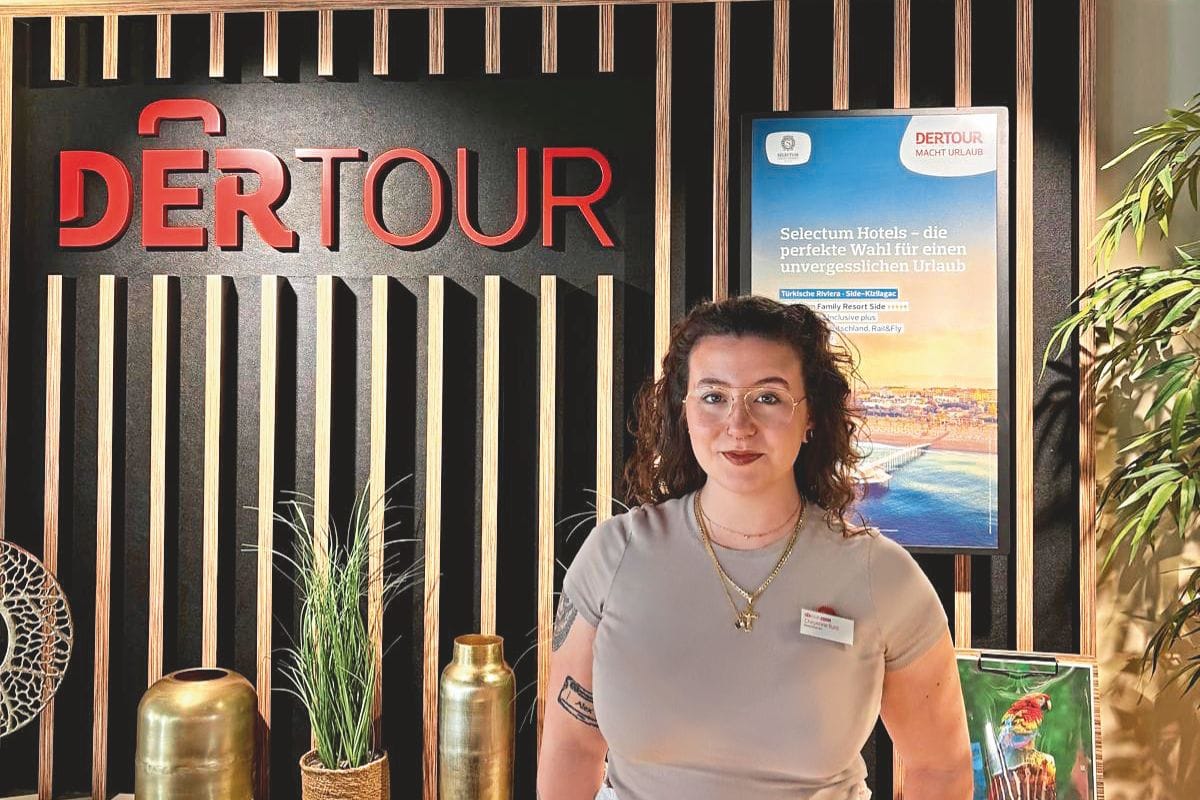 Eine junge Frau mit Brille und lockigem Haar steht vor einer Wand mit dem DERTOUR-Logo und einem Werbeplakat für Selectum Hotels in einem hellen Büro.