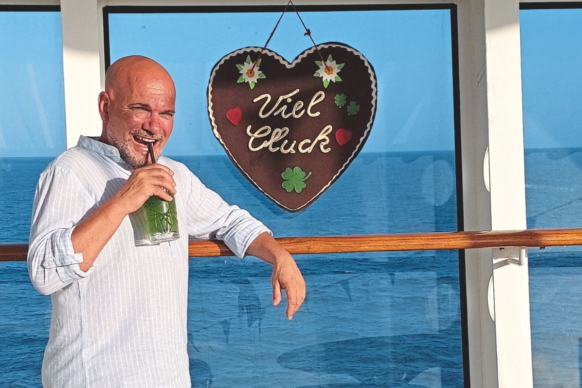 Ein glatzköpfiger Mann mit Bart trinkt aus einem Glas mit grünem Inhalt, während er an einem Geländer vor einer Glasscheibe mit Lebkuchenherz steht.