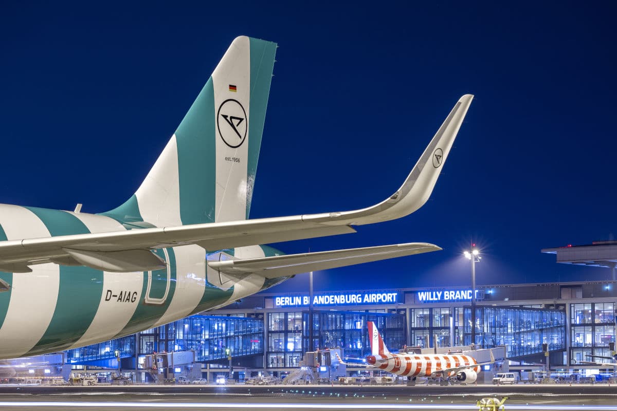 Das Heck eines Flugzeugs mit grün-weißem Streifenmuster steht auf dem Rollfeld vor dem beleuchteten Terminalgebäude des Berlin Brandenburg Airport bei Nacht.