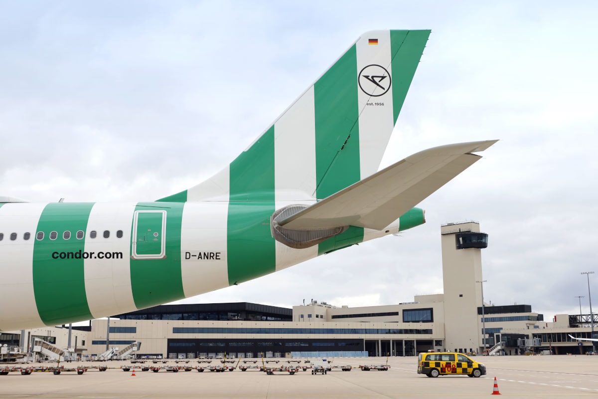 Das Heck eines Flugzeugs der Fluggesellschaft Condor mit grün-weißem Streifenmuster steht auf einem Rollfeld vor einem Flughafengebäude mit Kontrollturm.