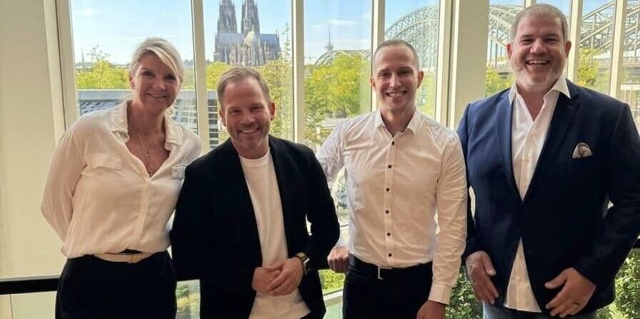 Vier Personen stehen lächelnd in einem hellen Raum vor einer großen Fensterfront, durch die der Kölner Dom und die Hohenzollernbrücke zu sehen sind.