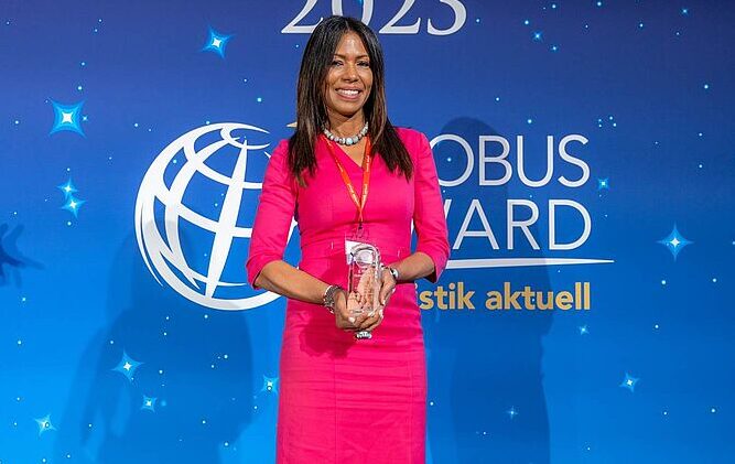 Eine Frau mit dunklen Haaren in einem pinkfarbenen Kleid hält eine gläserne Trophäe vor einem blauen Hintergrund mit Globus-Logo und Schriftzug Globus Award.