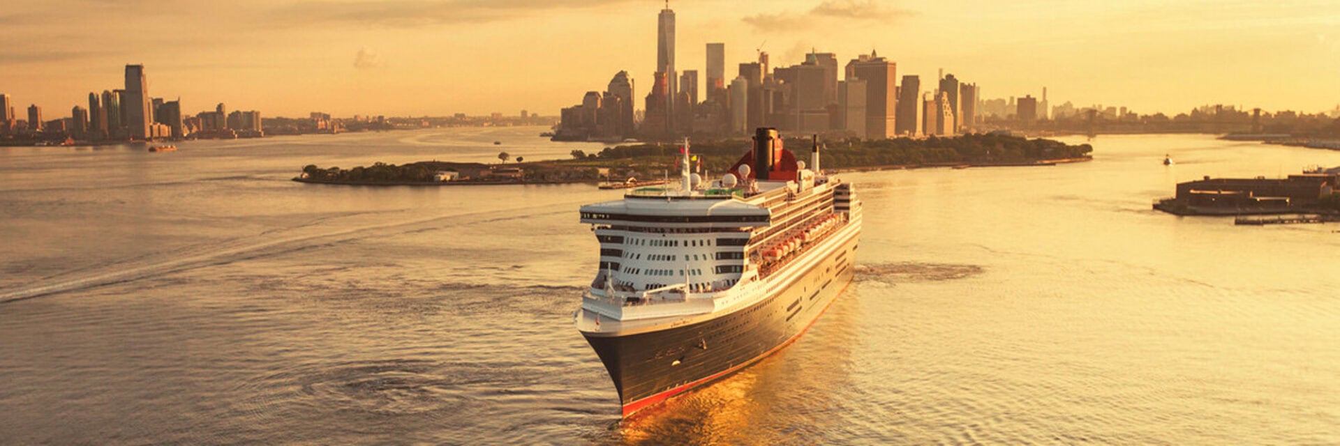 cunard-aufmacher Ein großes Kreuzfahrtschiff fährt bei warmem Sonnenuntergangslicht auf einem Fluss vor der Skyline von Manhattan in New York City in Richtung Betrachter.