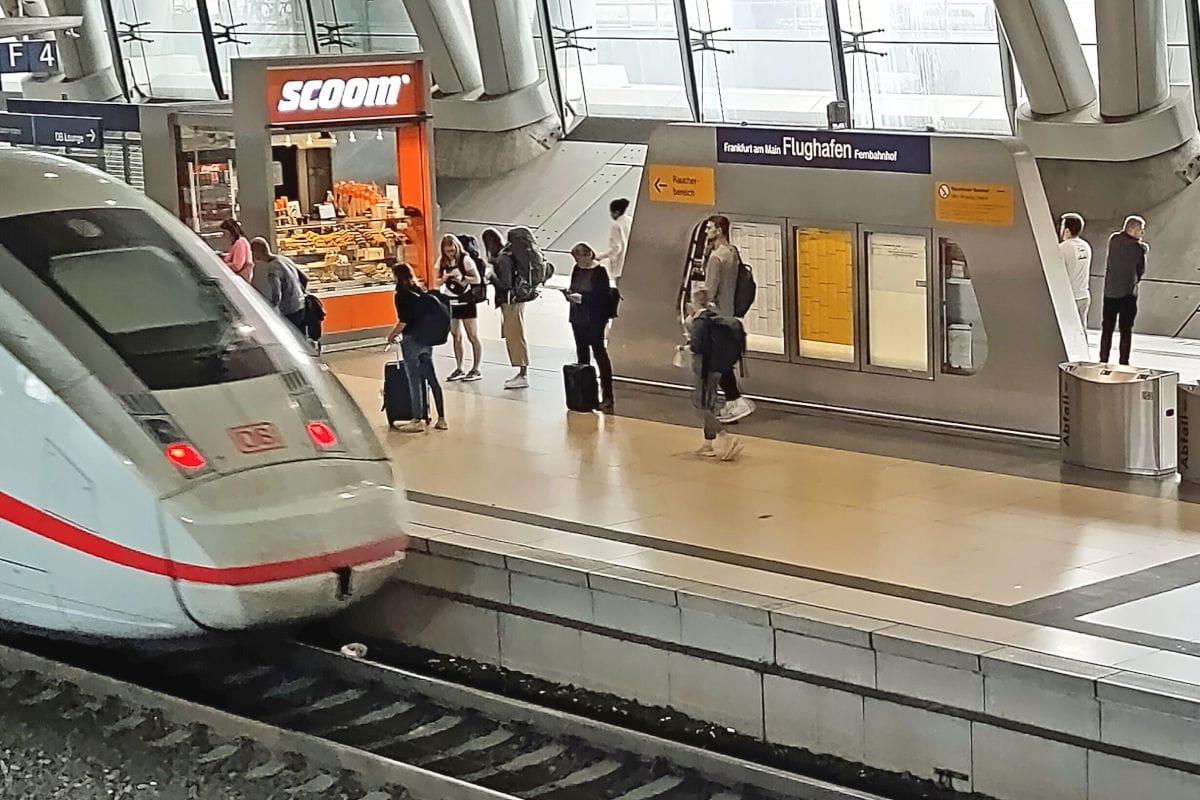 Ein ICE steht am Bahnsteig des Frankfurter Fernbahnhofs, während Reisende mit Gepäck vor einem Sccom-Verkaufsstand und Informationstafeln warten oder gehen.