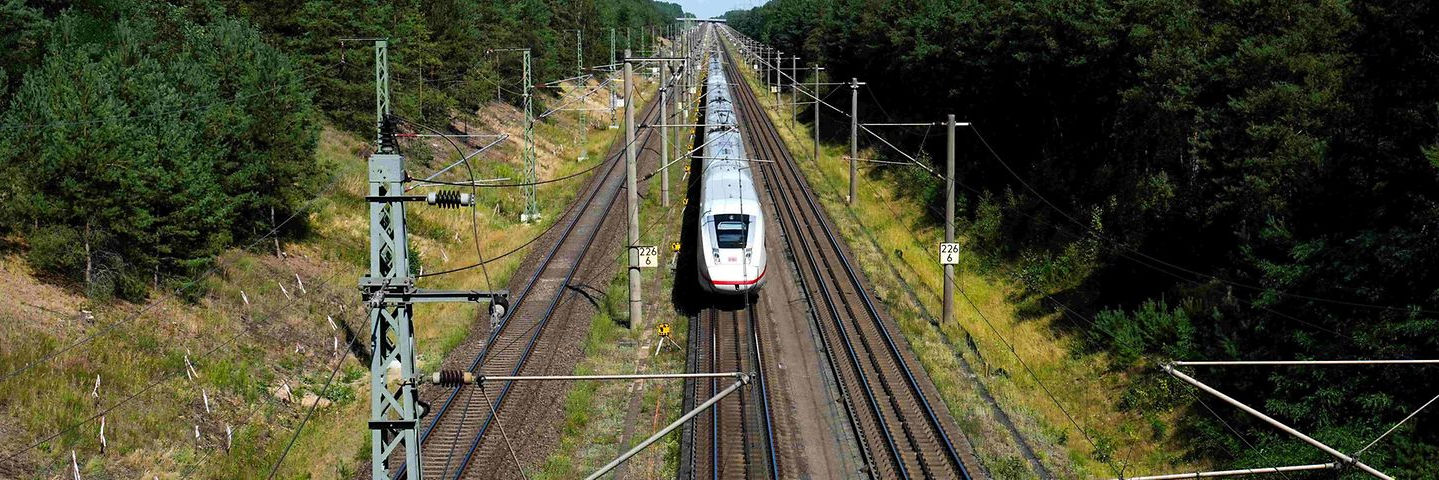 deutsche-bahn-ice-hannover-berlin Ein silberner ICE-Zug fährt auf einer zweigleisigen Bahnstrecke durch einen Wald, aufgenommen aus der Vogelperspektive mit Oberleitungen im Vordergrund.