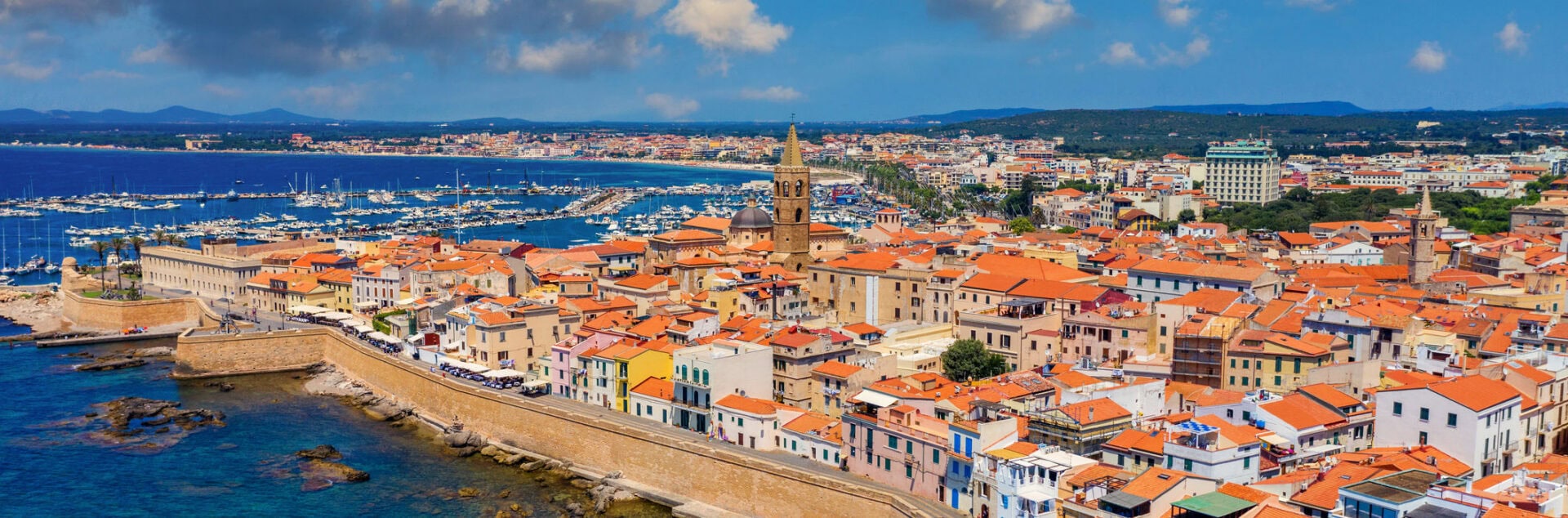 discover-airlines-italien Panoramablick auf die historische Altstadt von Alghero mit ihren roten Ziegeldächern, der Stadtmauer und dem angrenzenden Yachthafen unter blauem Himmel.