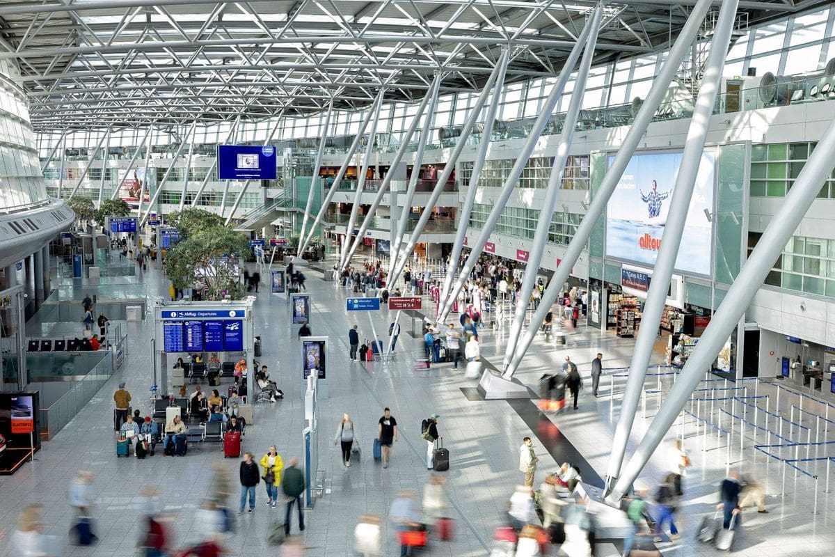 Weitwinkelaufnahme einer modernen Flughafenhalle mit hoher Glasdachkonstruktion, zahlreichen Reisenden, Anzeigetafeln und großen Werbeflächen an den Wänden.