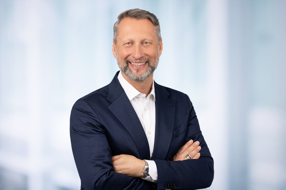Ein Mann mit grau meliertem Haar und Bart trägt ein weißes Hemd sowie ein dunkelblaues Sakko und lächelt vor einem hellen, unscharfen Hintergrund in die Kamera.