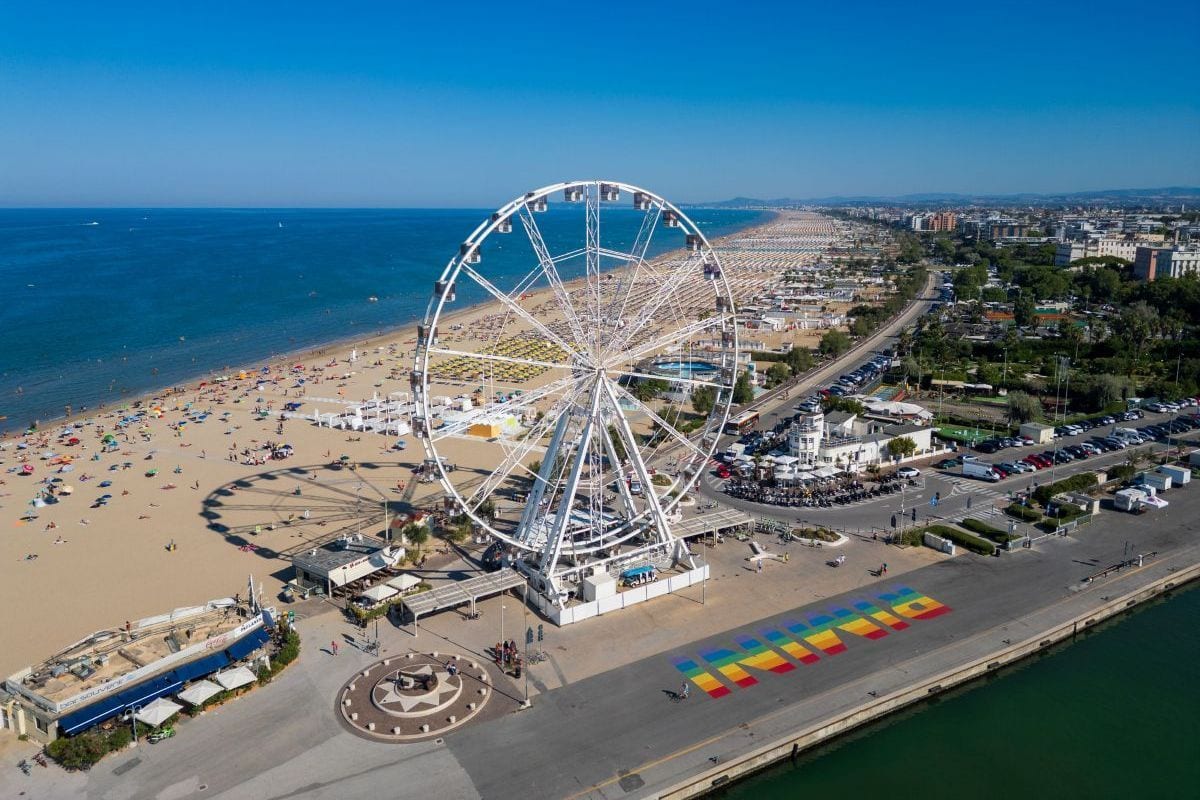 Luftaufnahme eines weißen Riesenrads an einem Sandstrand mit vielen Sonnenschirmen, angrenzender Küstenstraße und einem bunten Schriftzug auf dem Asphalt.