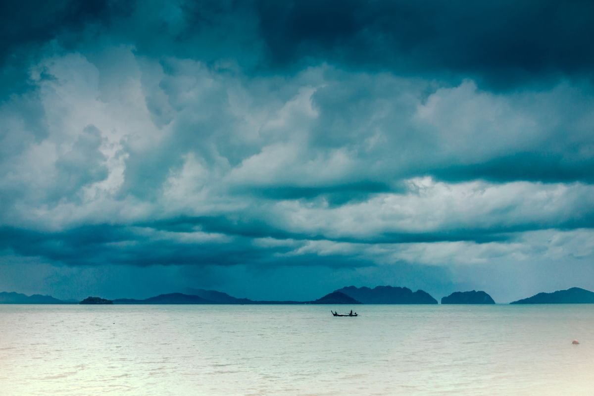 Ein kleines Boot auf dem ruhigen Meer vor einer Inselkette unter einem dramatischen, tiefblauen und wolkenverhangenen Himmel bei bewölktem Tageslicht.