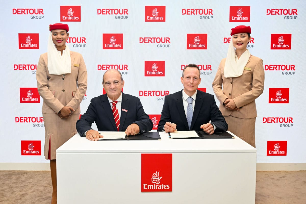 Zwei Männer sitzen an einem Tisch mit Verträgen, umgeben von zwei Flugbegleiterinnen von Emirates, vor einem Hintergrund mit Dertour-Logo.