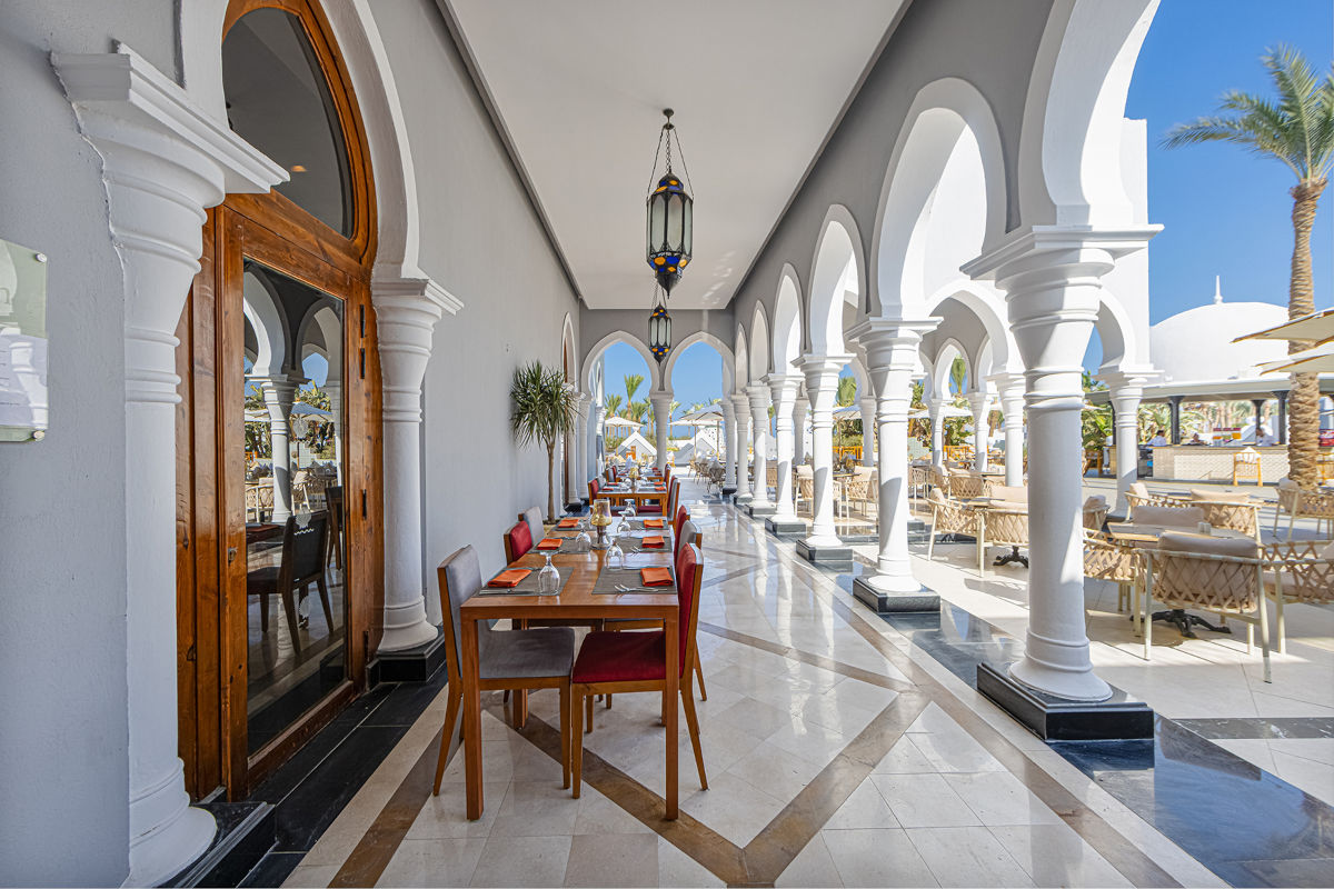 Eine überdachte Terrasse mit weißen Säulen und orientalischen Laternen, an der Holztische mit Stühlen in einer Reihe für Gäste bereitgestellt sind.