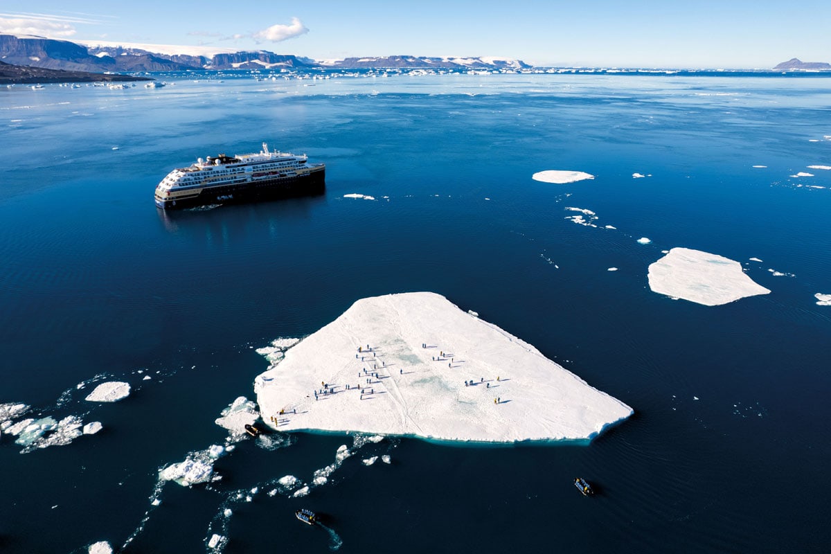Ein Kreuzfahrtschiff liegt in arktischen Gewässern vor einer großen Eisscholle, auf der zahlreiche Pinguine stehen, umgeben von weiteren kleinen Booten.
