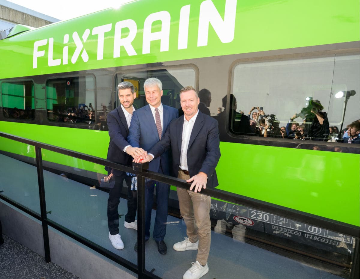 Drei Männer in Anzügen stehen vor einem leuchtend grünen Flixtrain-Waggon und drücken gemeinsam einen roten Knopf auf einem schwarzen Pult im Vordergrund.