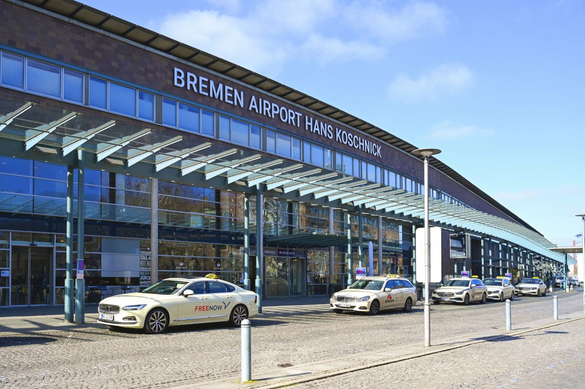 Die Fassade des Bremen Airport Hans Koschnick mit einer Glasfront und einem Vordach, davor stehen mehrere Taxis auf dem gepflasterten Vorplatz bei Tageslicht.