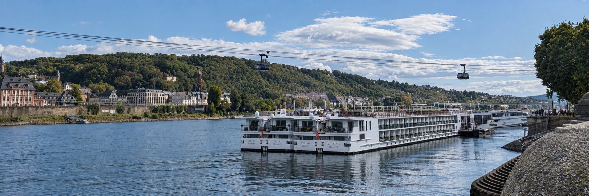 flusskreuzfahrt Ein weißes Flusskreuzfahrtschiff liegt am Ufer eines Flusses vor einer bewaldeten Hügellandschaft, während zwei Seilbahngondeln über das Wasser schweben.