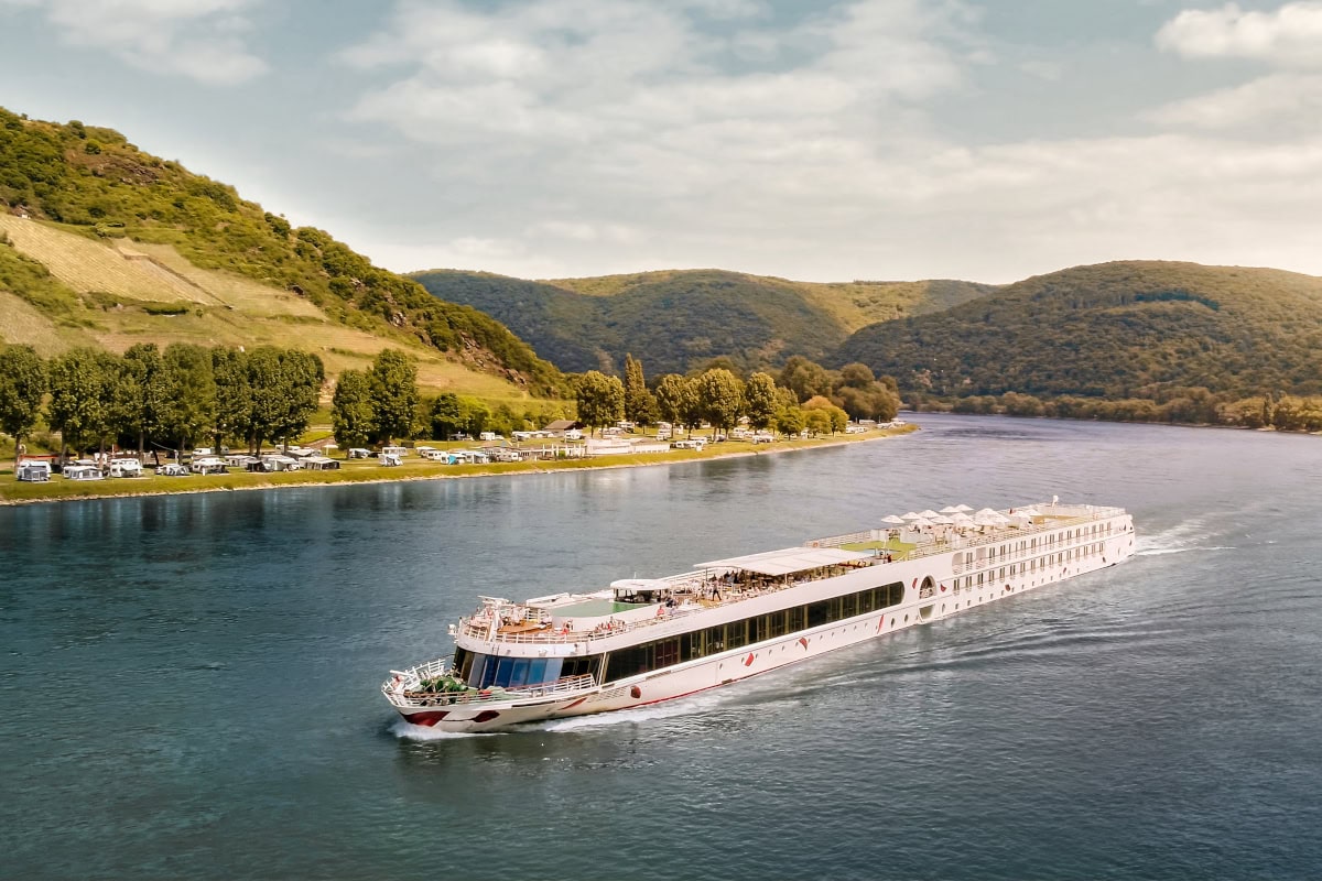 Ein weißes Flusskreuzfahrtschiff fährt auf einem breiten Fluss an einem grünen, hügeligen Ufer mit einem Campingplatz und Weinbergen bei Tageslicht vorbei.