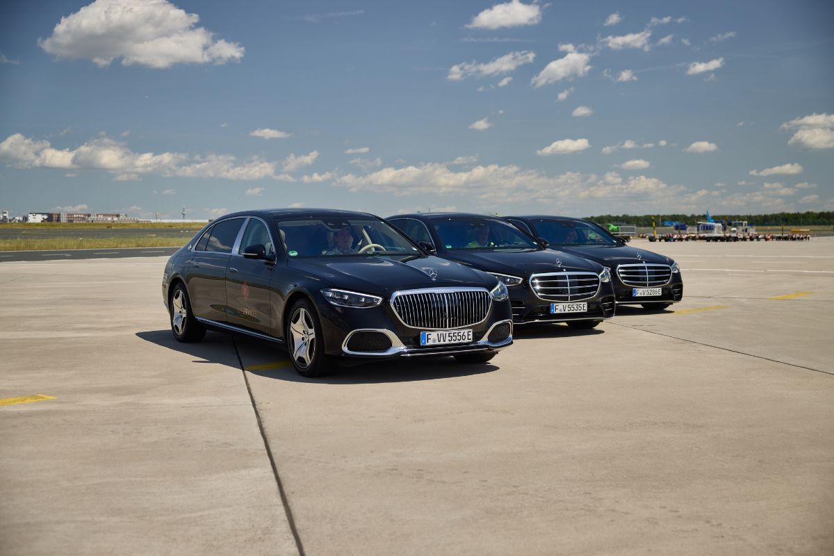 Drei schwarze Mercedes-Benz Limousinen stehen hintereinander auf einer asphaltierten Fläche vor einem blauen Himmel mit weißen Wolken an einem sonnigen Tag.