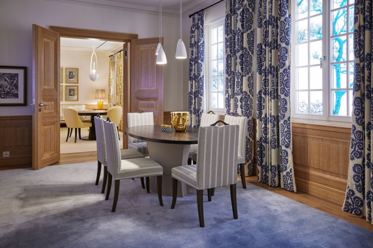 Ein eleganter Essbereich mit einem runden Tisch, gestreiften Stühlen und blauen Vorhängen, der durch eine offene Holztür in einen weiteren Raum führt.