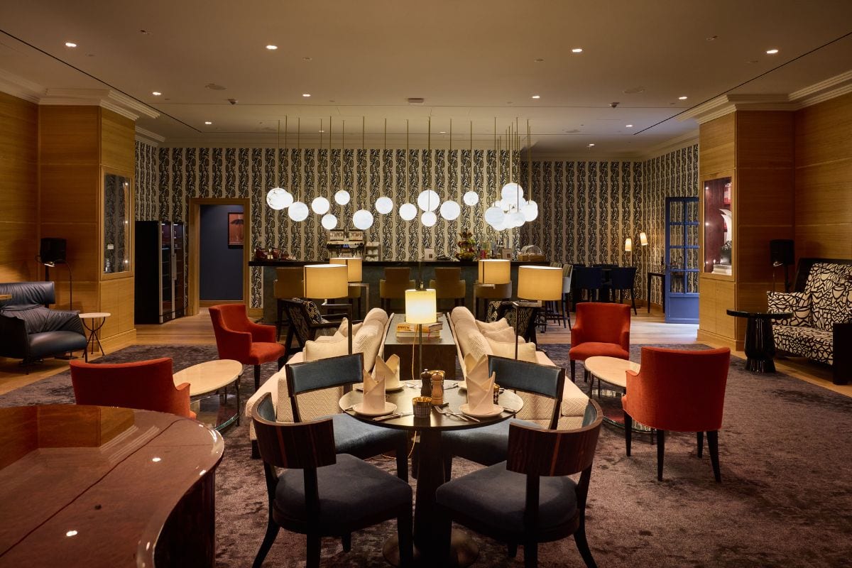 Ein eleganter Hotel-Loungebereich mit runden Tischen, gepolsterten Stühlen, einer Bar im Hintergrund und einer dekorativen Kugel-Lichterkette an der Decke.