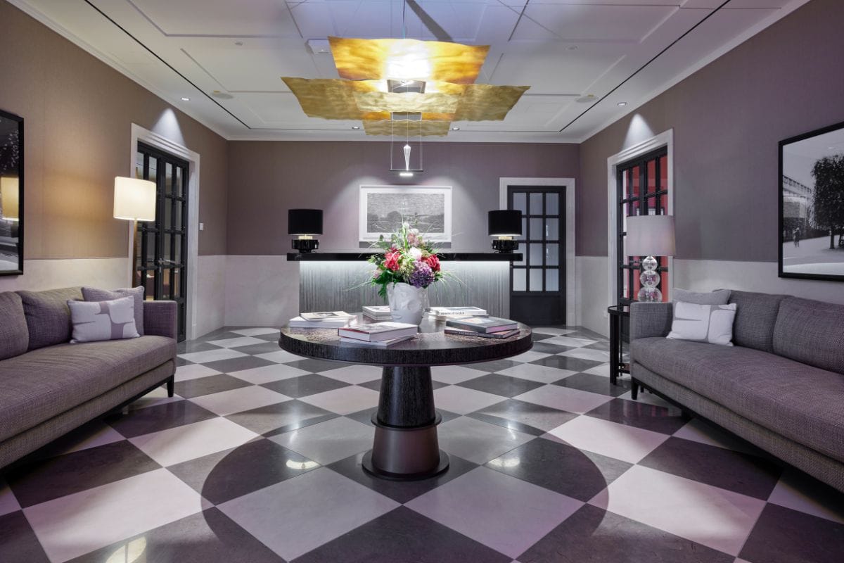 Ein eleganter Hotel-Empfangsbereich mit kariertem Boden, zwei grauen Sofas, einem runden Tisch mit Blumenstrauß und einer zentralen, goldenen Deckenlampe.