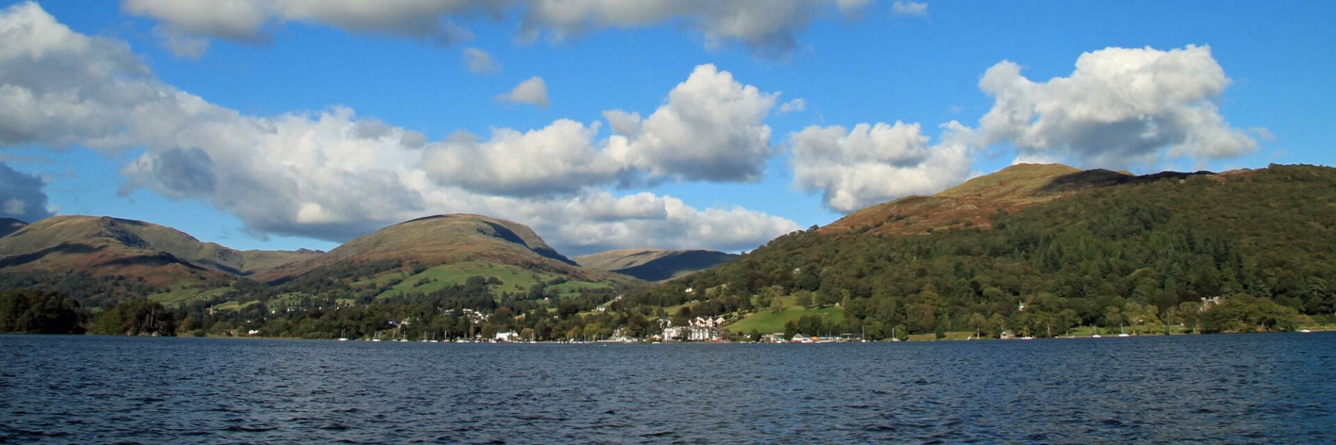 Panoramablick auf einen See mit bewaldeten Hügeln im Hintergrund unter einem blauen Himmel mit weißen Wolken, am Ufer sind kleine Gebäude zu erkennen.