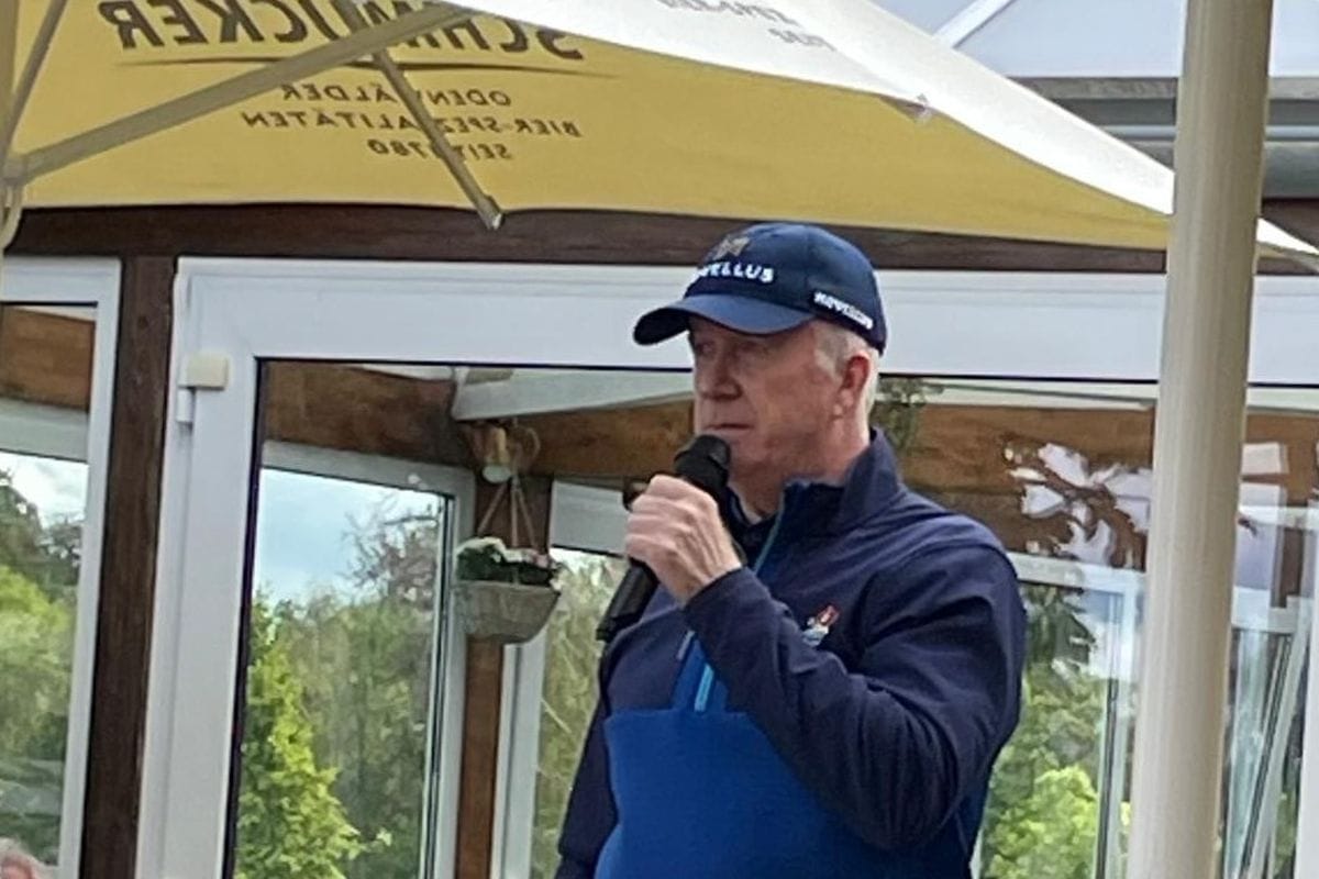 Ein Mann mit blauer Baseballkappe und dunkler Jacke spricht in ein Mikrofon, im Hintergrund ist ein gelber Sonnenschirm mit Schmucker-Logo zu sehen.