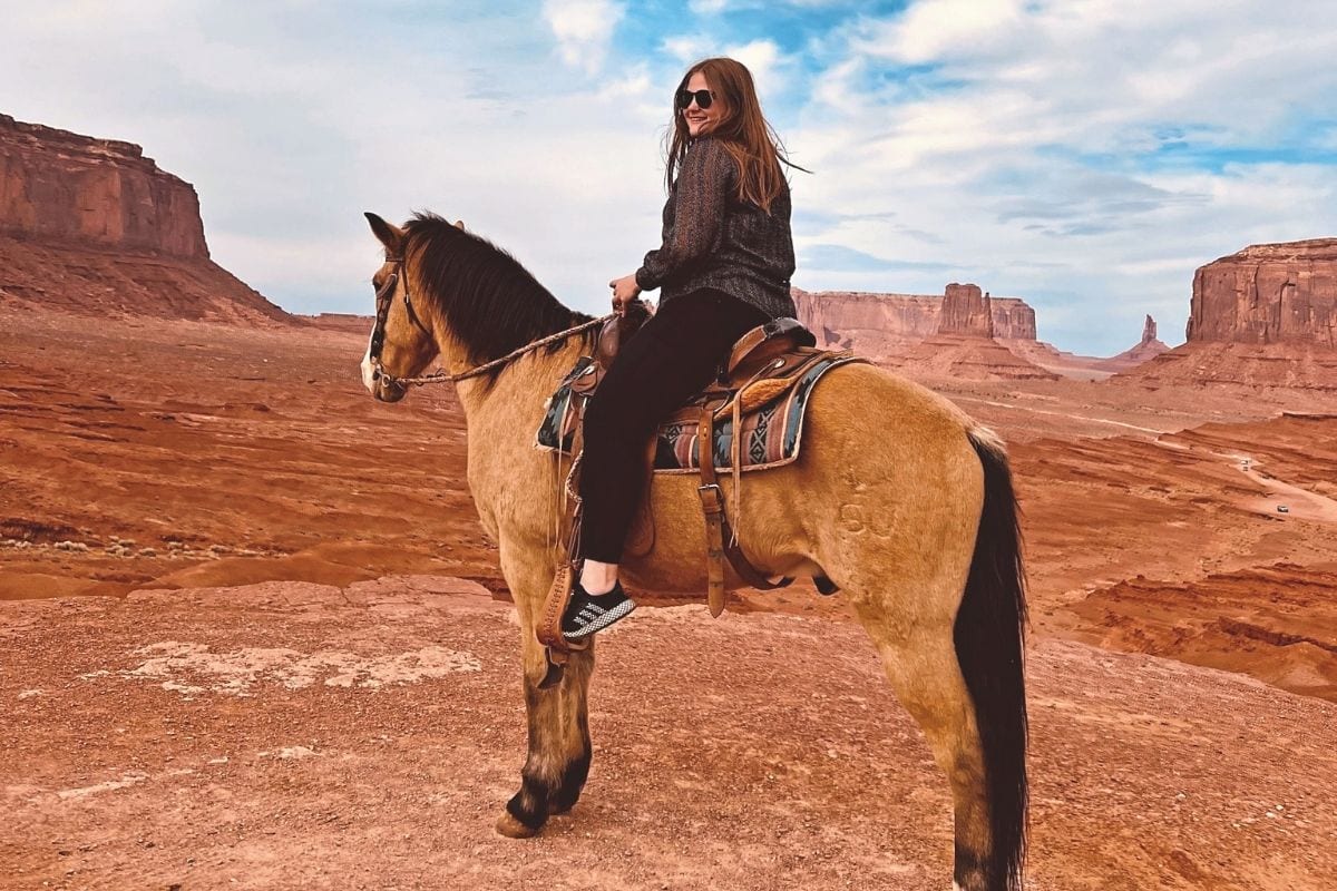 Eine junge Frau mit Sonnenbrille reitet auf einem hellbraunen Pferd durch eine weite, rote Wüstenlandschaft mit markanten Felsformationen im Hintergrund.