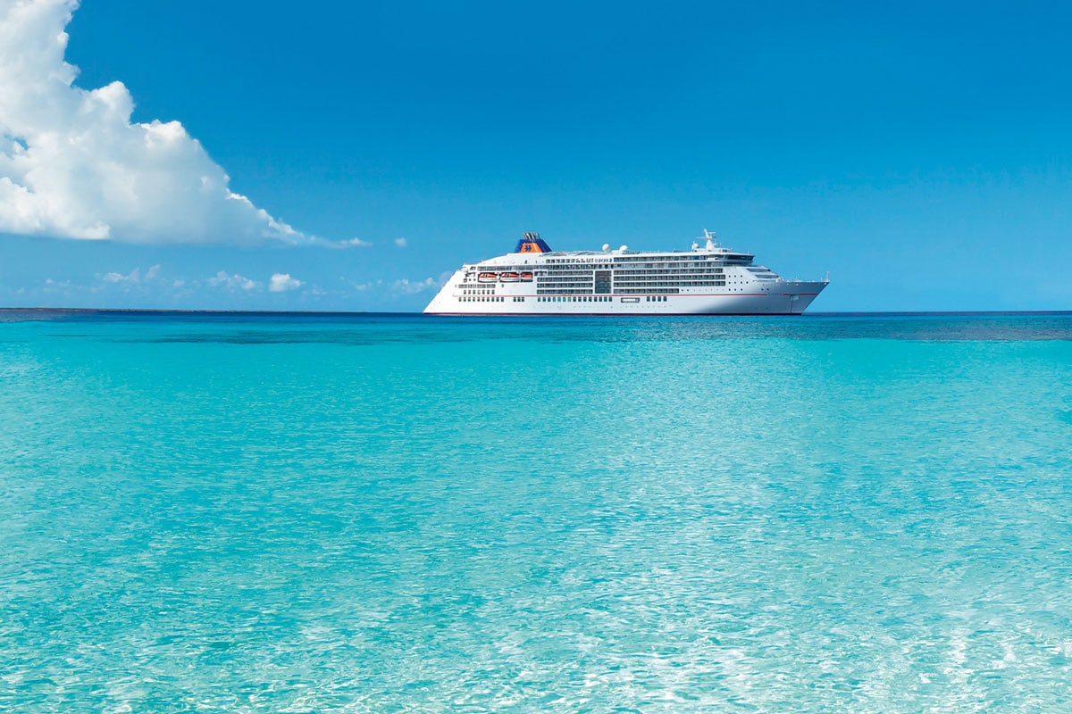 Ein weißes Kreuzfahrtschiff der Reederei Hapag-Lloyd Cruises fährt auf dem klaren, türkisfarbenen Wasser unter einem strahlend blauen Himmel mit Wolken.