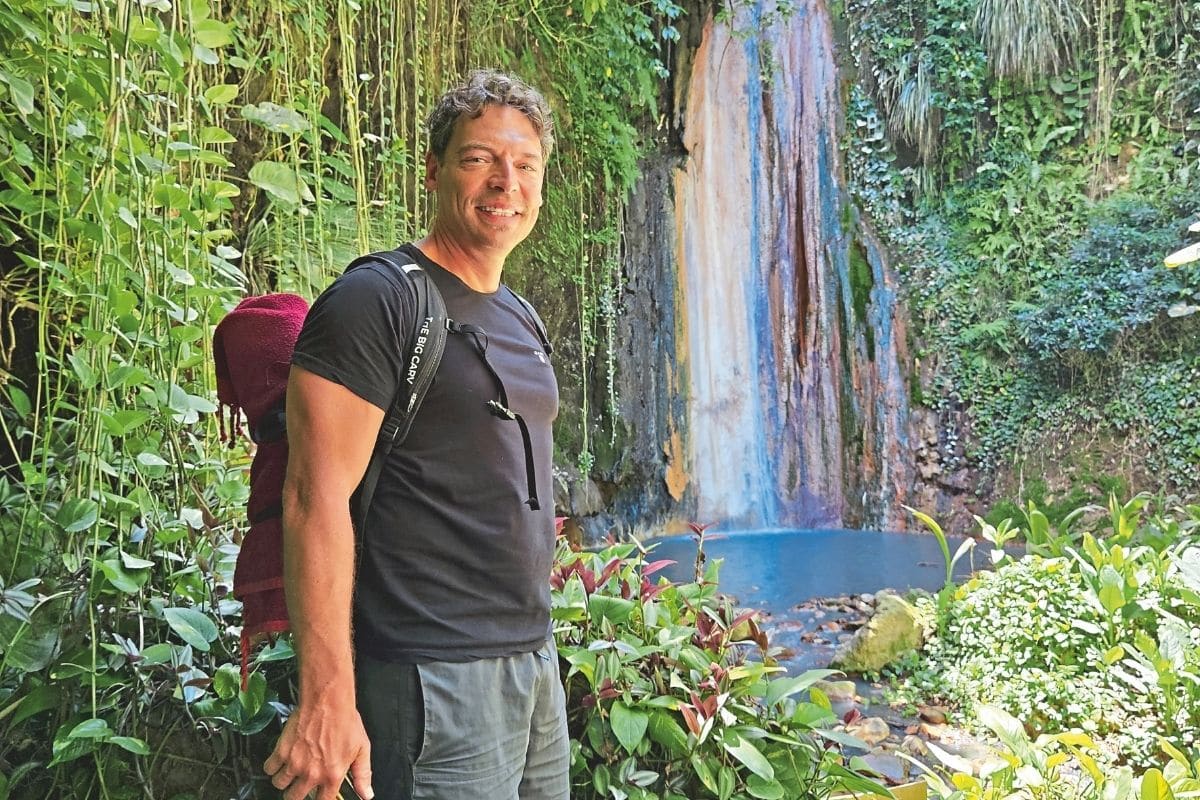 Ein lächelnder Mann mit Rucksack steht vor einem kleinen Wasserfall in einer üppigen, grünen Felsnische mit dichtem Pflanzenbewuchs und einem Wasserbecken.