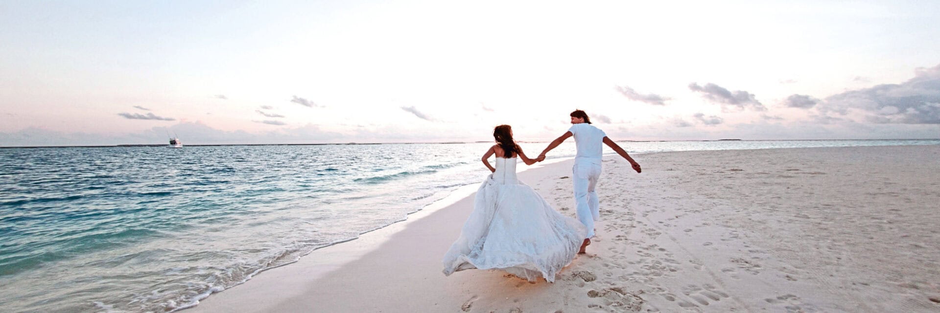 hochzeitsreisen-beratung Ein Paar in weißer Kleidung läuft Hand in Hand am hellen Sandstrand entlang, während im Hintergrund das Meer und ein heller Himmel zu sehen sind.