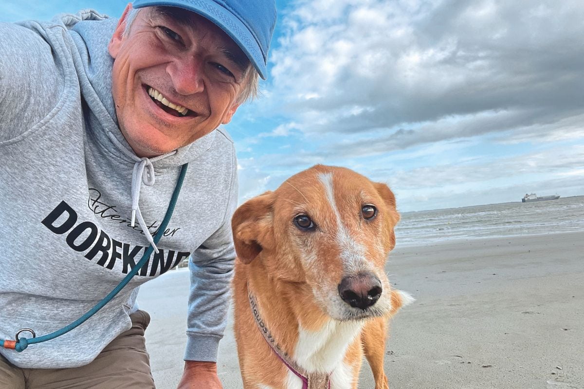 Ein lächelnder Mann mit blauer Kappe und grauem Kapuzenpullover macht ein Selfie mit einem braun-weißen Hund an einem weitläufigen Sandstrand bei Bewölkung.