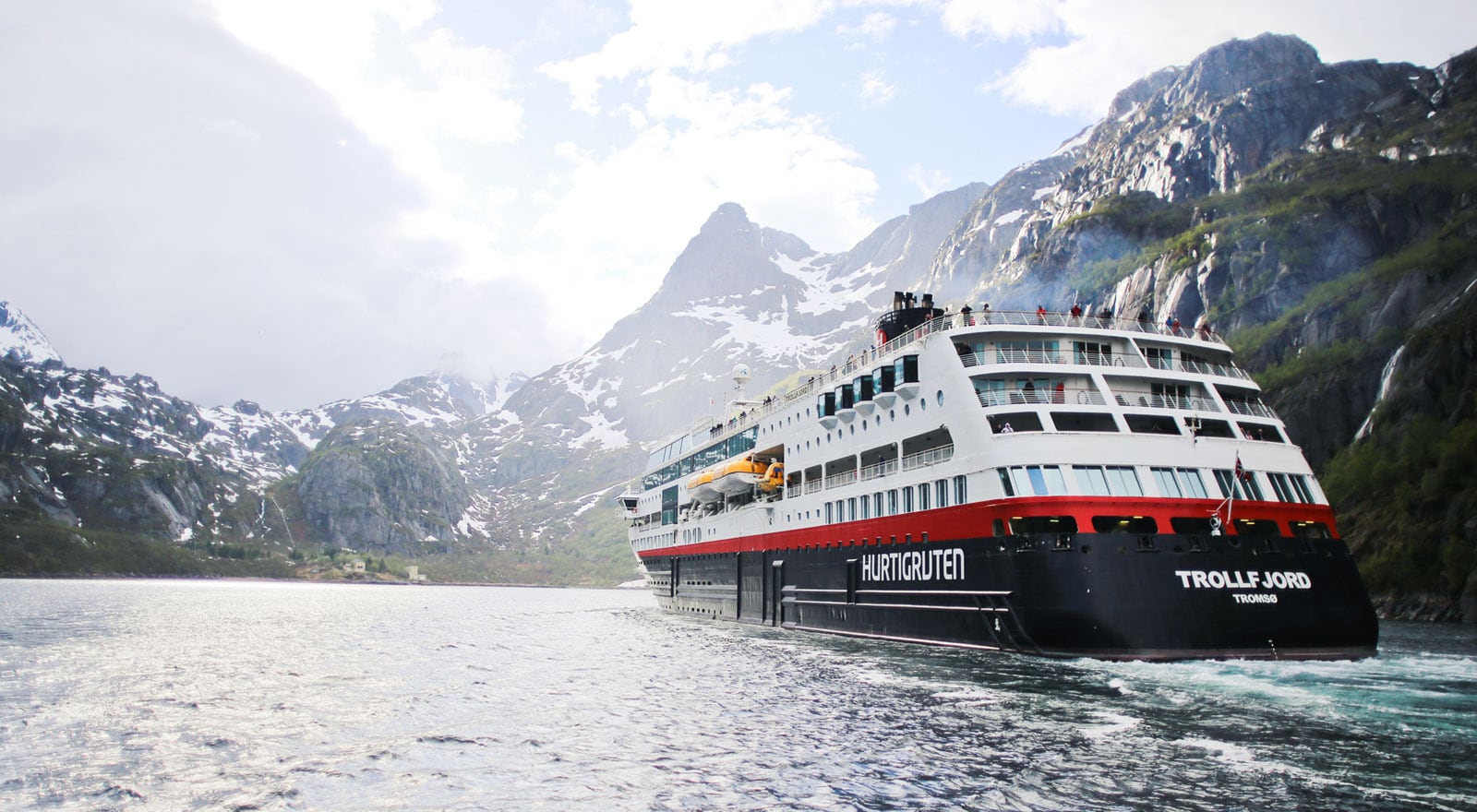 Das Hurtigruten-Schiff Trollfjord fährt durch einen norwegischen Fjord, umgeben von hohen, schneebedeckten Bergen unter einem hellen, bewölkten Himmel.