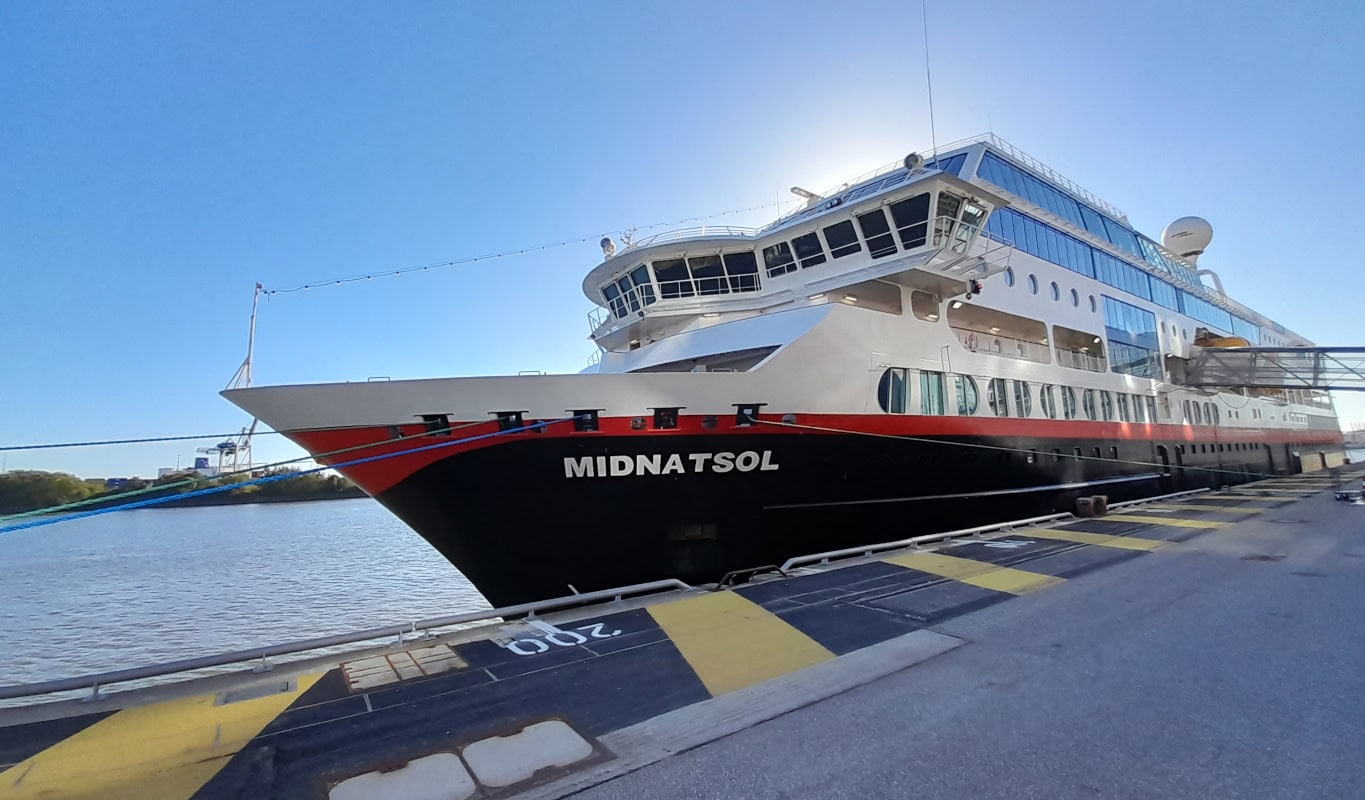 Die Midnatsol liegt an einer Kaimauer mit gelb-schwarzer Markierung vor Anker; das Schiff ist weiß und schwarz mit rotem Zierstreifen am Rumpf lackiert.