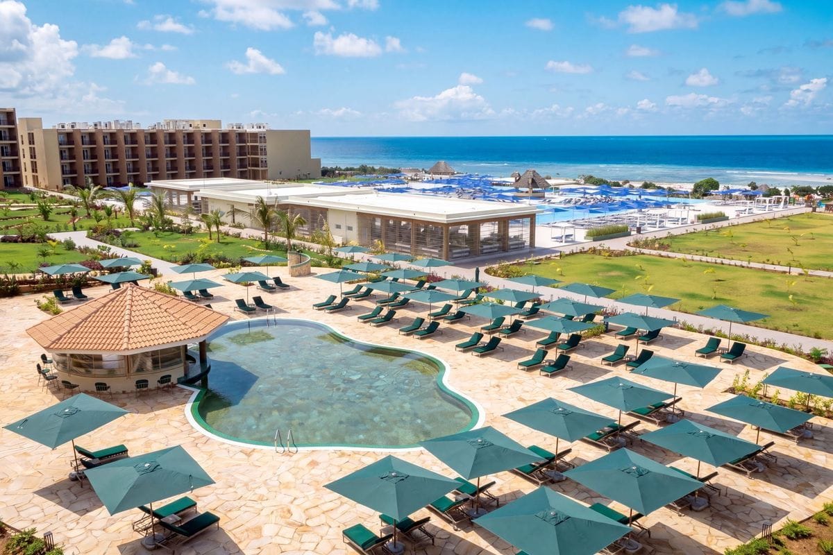 Blick von oben auf eine Hotelanlage mit geschwungenem Pool, zahlreichen grünen Sonnenschirmen, Liegen auf einer Steinterrasse und dem blauen Meer im Hintergrund.