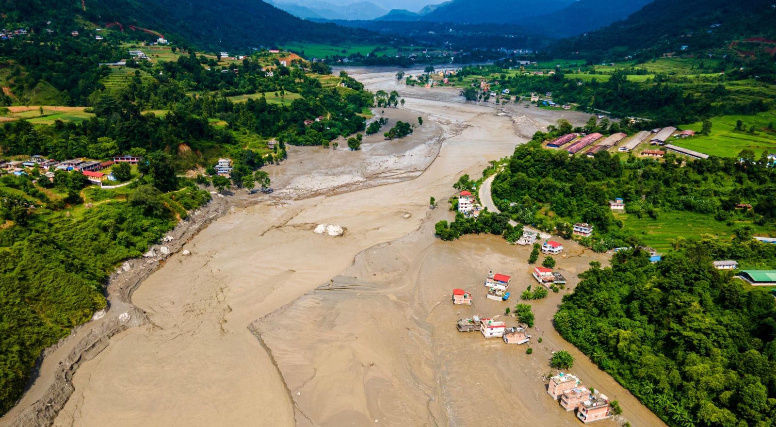 Luftaufnahme einer von Schlamm und Sedimenten überfluteten Flusslandschaft in einem grünen Tal, umgeben von bewaldeten Hügeln und verstreuten Siedlungen.