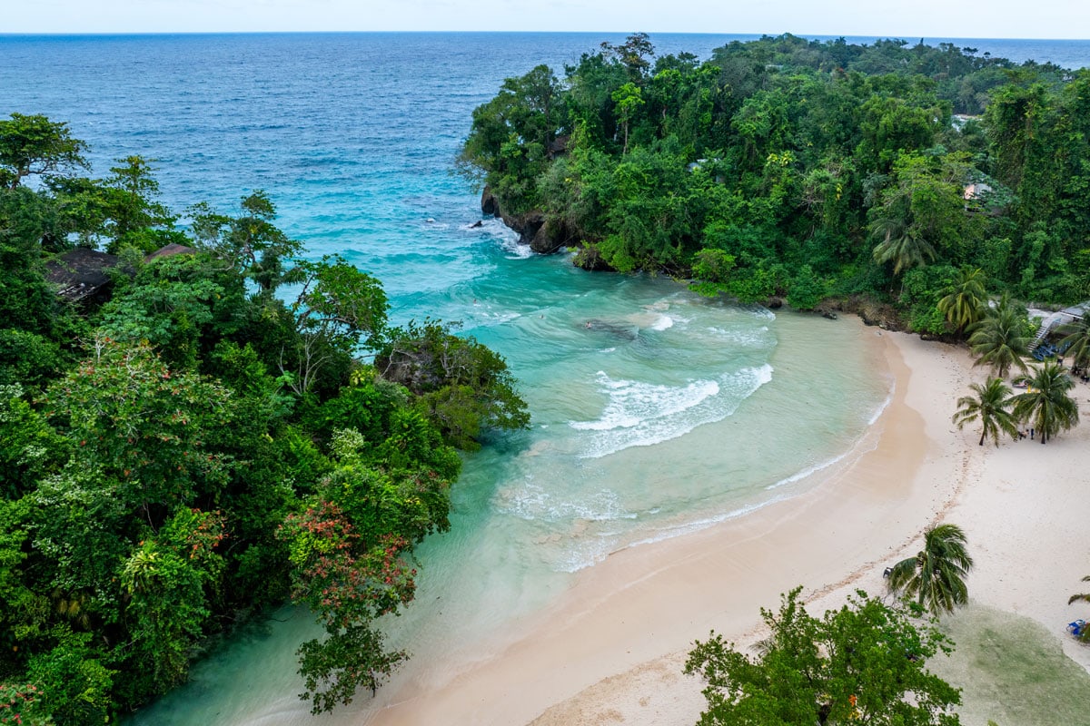 Luftaufnahme einer tropischen Bucht mit hellem Sandstrand, umgeben von dichtem, grünem Wald und türkisblauem Wasser, das sanft an das Ufer heranrollt.