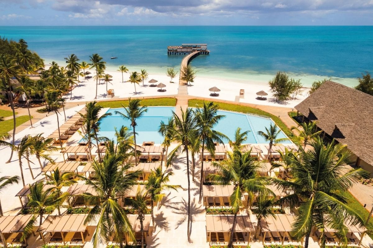 Luftaufnahme eines Resorts mit Poollandschaft, Palmen, weißem Sandstrand und einem langen Steg, der in das türkisfarbene Meer führt, unter blauem Himmel.