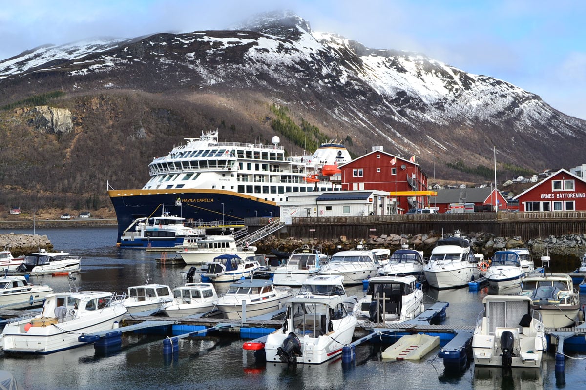 Ein Hafen mit vielen Booten, einem großen Schiff namens Havila Capella und roten Gebäuden im Hintergrund, umgeben von Bergen.
