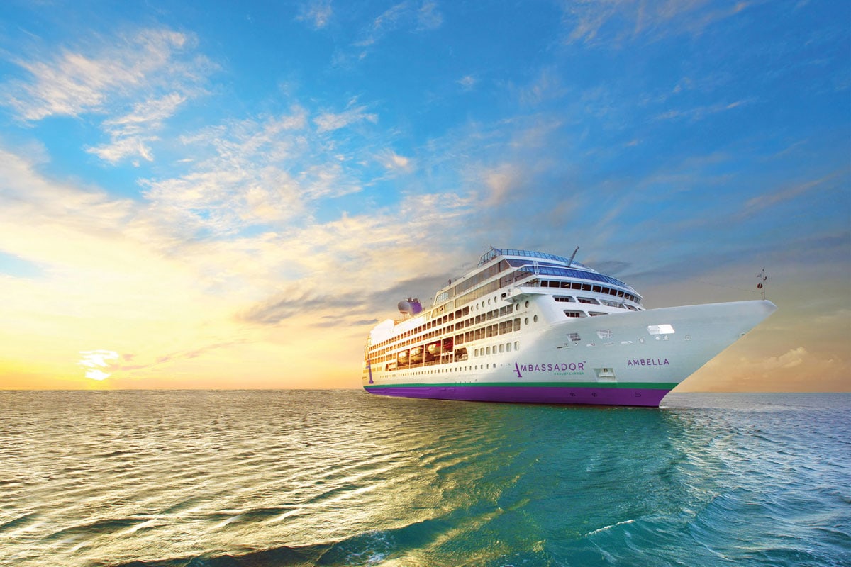 Das Kreuzfahrtschiff Ambella der Reederei Ambassador fährt bei Sonnenuntergang auf dem offenen Meer unter einem wolkenverhangenen, hellen Abendhimmel.