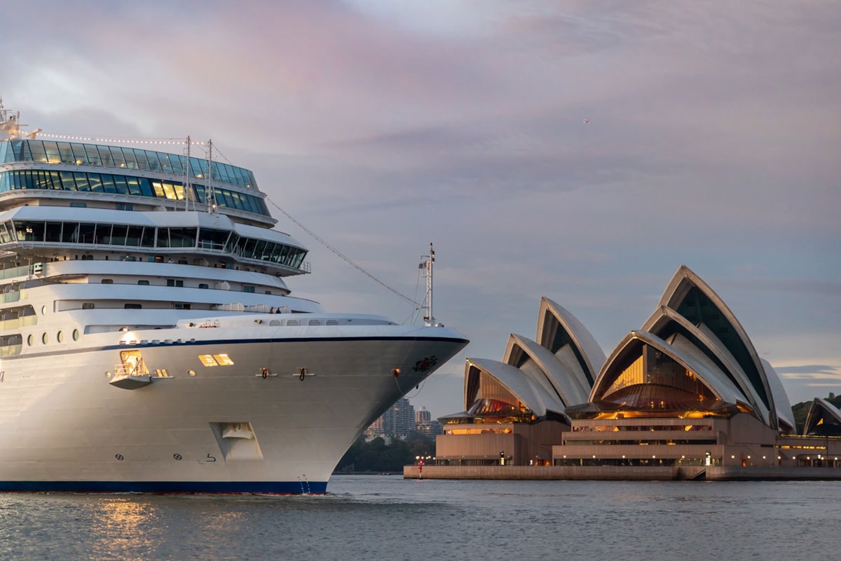 Ein großes Kreuzfahrtschiff fährt an der Sydney Opera House vorbei, das im Abendlicht beleuchtet ist.